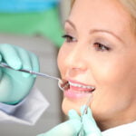 A close-up of a dental patient, a woman with blonde hair, undergoing a dental examination or procedure. She is smiling and appears calm as a dentist wearing teal gloves holds a dental mirror and examines her mouth. The bright light of the dental office shines on her face, emphasizing the cleanliness and precision of the dental care environment.