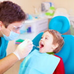 A young boy sitting in a blue dental chair with his mouth open, being examined by a dentist. The dentist, wearing protective glasses, a mask, gloves, and a blue scrubs, is using a dental mirror to check the boy's teeth. The boy is wearing a red shirt and a blue bib, appearing calm and cooperative. The background shows a clean and organized dental clinic with various dental tools and equipment.