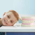 A young boy with short blond hair smiling and resting his head on a white table in a dental office. Next to him is a large dental model showing a set of teeth and gums. The boy is wearing a blue plaid shirt with a gray hood, and the background features a light blue wall with dental equipment, creating a cheerful and educational atmosphere.