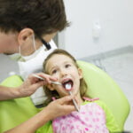 A young girl in a pink polka-dot apron and green shirt sitting in a dentist's chair with her mouth open. A dentist, wearing a mask and glasses, is examining her teeth using a dental mirror and probe. The girl appears calm and cooperative. The setting is a modern dental clinic with equipment and tools visible in the background.