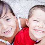 Two young children smiling brightly at the camera. The girl on the left has short brown hair with a hint of purple, wearing a white top, and has a few missing teeth. The boy on the right, in a red shirt, has short brown hair. They are close together, and the girl has her arm around the boy's shoulder. The background is a blurred outdoor setting, possibly stone steps.