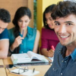 A cheerful young man with curly dark hair and a denim shirt smiling at the camera while three other people are engaged in studying or working in the background. The group, seated at a table, includes two women and one man, all focused on their books and notes. The setting appears to be a casual and collaborative learning environment.
