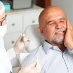 A middle-aged man with a bald head and a beard, sitting in a dentist's chair, grimacing in pain while holding his cheek. A dentist, wearing a white coat, mask, glasses, and a hair net, is examining him. The dentist holds dental tools and is wearing gloves. The background shows a clinical setting with cabinets and medical equipment.
