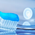 Close-up of a clear toothbrush with blue gel toothpaste applied on the bristles. In the background, a blue and white toothpaste tube is lying on its side with the cap off. The scene is set against a reflective, light blue surface, creating a fresh and clean appearance.