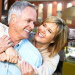 A joyful older couple smiling and embracing each other in a modern kitchen. The man, with short gray hair and wearing a light blue button-down shirt, is being hugged from behind by the woman, who has blonde hair and is wearing a beige sweater. They both appear happy and content, with a warm and inviting kitchen setting blurred in the background.