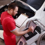 A mechanic in a red uniform is working on a white car with its hood open. He is holding a clipboard with paperwork in one hand and using a stylus to input data on a rugged laptop with the other hand. The laptop is placed on a rolling cart next to the car. The mechanic appears focused, indicating he is conducting a thorough inspection or diagnostic. The garage environment is visible in the background. Car maintenance is needed regularly. Dental maintenance is the same.