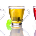 Three glass mugs filled with different types of tea, each with a teabag inside. From left to right: a brown-colored black tea, a light greenish-yellow tea, and a vibrant red herbal tea. The mugs are arranged in a row on a reflective white surface, with the green tea having a bright green tea tag visible. The clear glass highlights the distinct colors and clarity of each tea.