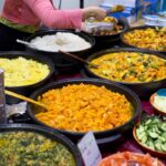 A variety of colorful Indian dishes displayed in large pans on a buffet table illustrates the wide variety of flavors that are recognized by the tongue. Visible dishes include yellow rice, white rice, various curries with vegetables, and fresh salads with sliced cucumbers and chopped greens. A person in a pink shirt and apron is serving food into a takeaway container. Foil trays for takeout are stacked on the side. The background shows more food items and containers, suggesting a bustling food market or festival setting.
