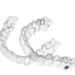 A pair of clear dental aligners placed on a white background. These transparent orthodontic devices are designed to fit over the teeth and gradually straighten them. The aligners have intricate detailing to match the contours of the teeth.