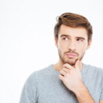 A contemplative young man with styled hair and a light beard is pictured against a white background, his hand resting thoughtfully on his chin. He wears a casual grey T-shirt and his gaze is directed upwards, suggesting he is deep in thought or consideration.