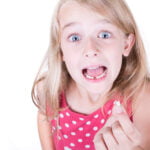 A young girl with blonde hair and wide blue eyes is wearing a pink polka dot dress. She has an excited expression, mouth open, and is holding a small tooth in her hand, showing the gap from where it fell out. The background is plain white.