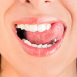 Close-up of a woman's smile, showcasing her white teeth and a tongue piercing with a metallic ball. The focus on her open mouth with glossy lips emphasizes oral jewelry and dental aesthetics. The background is blurred to highlight the piercing as the central element of the image.