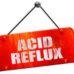 A red, rectangular wooden sign hanging by a black and white rope, with the words "ACID REFLUX" written in bold white letters. The sign has a distressed, weathered look.