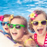 Three young girls wearing brightly colored sunglasses and flower hair accessories, smiling and leaning on the edge of a swimming pool. The girls are dressed in swimsuits and enjoying a sunny day in the water.