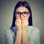 A young woman with long brown hair, wearing black-framed glasses and a light blue shirt, looks nervously at the camera. She holds her hands close to her mouth, covering part of her face, and her wide eyes express anxiety. The background is a plain, muted gray.