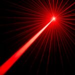A bright red laser beam emanates from a single point, creating an array of radiating lines against a black background. The intense central point of the laser is surrounded by multiple thin, straight beams that spread outward in all directions, creating a striking and dramatic visual effect.