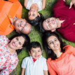 A family of six, comprising three men and three women, lies on their backs in a circle on the grass, heads touching in the center. They are all smiling and looking up at the camera. The adults wear colorful clothes: the older man in an orange striped polo, the older woman in a floral blouse, a young man in a red shirt, and a young woman in an orange top. Two children, a boy in a white shirt and a girl in a white dress, complete the circle, creating a warm, joyful family portrait.