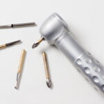 A close-up of a dental drill with a textured silver handle and various drill bits laid out on a white surface. The drill bits, made of metal and appearing in different shapes and sizes, are designed for dental procedures, showcasing the tools used in dentistry for precision work.