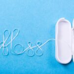 A white dental floss container with its lid open, placed on a light blue background. The floss extends from the container and is artistically arranged to spell the word "floss