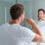 A man in a white T-shirt is brushing his teeth while looking at his reflection in a bathroom mirror. The image is taken from over his shoulder, creating a perspective where his focused expression is visible as he engages in his oral hygiene routine. The bathroom appears modern and well-lit, suggesting a clean and healthy lifestyle.