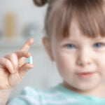 A young girl with light brown hair and bangs, wearing a striped shirt, holds a small blue pill between her fingers. She is smiling softly, and the background is blurred, focusing attention on her and the pill. The image conveys a sense of trust and care in a medical or health-related context.