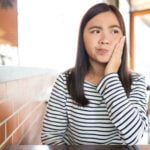 A young woman with long dark hair, wearing a striped shirt, sits at a table in a brightly lit cafe. She appears to be experiencing discomfort, holding her right cheek with a pained expression on her face. The setting suggests she might be dealing with a toothache or similar dental issue.