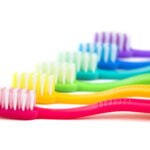 A row of colorful toothbrushes is lined up diagonally on a white background. Each toothbrush has a different vibrant color, including pink, yellow, green, blue, and purple. The bristles on the toothbrushes are white, with some having color accents that match the handle. The image focuses on the bright and varied designs of the toothbrushes, emphasizing dental hygiene.