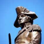 A bronze statue of a historical figure wearing a tricorne hat and military uniform. The figure has a serious expression and detailed epaulets on the shoulders. The statue is set against a clear blue sky, highlighting the craftsmanship and features of the monument.
