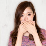 A young woman with long brown hair is covering her mouth with both hands, expressing a look of surprise or concern. She is wearing a colorful, textured top and stands against a light wooden background. Her eyes are wide open, and her facial expression suggests she might be reacting to something shocking or unexpected such as bad breath.