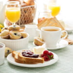 A breakfast spread featuring a variety of items on a light green tablecloth. In the foreground, a plate with toast topped with berry jam, a small pat of butter, and fresh raspberries is accompanied by a cup of coffee. Surrounding items include a bowl of blueberry muffins, a dish of mixed berries, a glass of orange juice, a basket of bread, and a pitcher of milk. The setting is bright and inviting, with natural light illuminating the food.