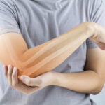 A person wearing a gray shirt is holding their left elbow with their right hand, indicating discomfort or pain. The image includes a semi-transparent overlay of the arm's bone structure, showing the humerus, radius, and ulna, to emphasize the skeletal anatomy. The background is softly blurred, keeping the focus on the arm and the highlighted bones.