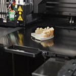 A 3D printer in action, creating a dental model. The model, resembling human teeth and gums, is partially completed and sits on the printer's build platform. The printer's mechanical and electronic components, with warning labels, are visible in the background. The scene is set in a modern dental lab, showcasing the use of advanced technology in dental prosthetics and orthodontics.