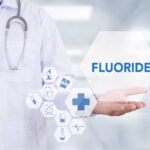 A medical professional wearing a white lab coat and a stethoscope around their neck is presenting the word "FLUORIDE" in blue text on a transparent hexagonal shape. Surrounding the word are various medical icons, including a microscope, syringe, stethoscope, test tube, and medical cross. The background is a blurred, sterile environment, likely a hospital or clinic.