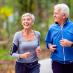 An older couple is jogging together on a wooded trail. The woman, with short gray hair, wears a gray long-sleeve top and black athletic pants, smiling brightly with earphones and a smartphone armband. The man, with white hair, is dressed in a blue zip-up jacket and dark pants, also smiling as they enjoy their run. The background is filled with green trees and foliage, suggesting a peaceful, outdoor setting.