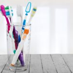 A glass filled with various colorful toothbrushes is placed on a light wooden surface. The toothbrushes have different handle designs and colors, including purple, green, blue, and orange. The background is softly lit with white light, creating a clean and bright atmosphere.