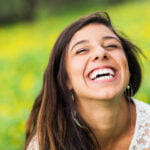 A young woman with long brown hair is laughing joyfully in an outdoor setting. She is wearing a white lace top and hoop earrings. The background is a blurred field of green grass and yellow flowers, suggesting a sunny, vibrant day. Her expression is one of pure happiness, with her eyes closed and mouth wide open in a carefree laugh.