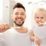 A smiling man and a young girl are brushing their teeth together in a brightly lit bathroom. The man has short brown hair and a beard, holding a green toothbrush. The girl, with blonde hair tied back, is using a white toothbrush. Both are wearing white shirts and appear happy, sharing a moment of dental hygiene. The background shows bathroom fixtures and a mirror
