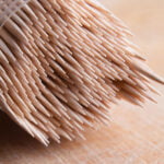 A close-up view of a clear plastic container filled with wooden toothpicks. The toothpicks are densely packed with their pointed ends facing outward, creating a pattern of sharp tips. The background is a soft, textured surface, slightly out of focus.