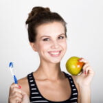 A young woman with a cheerful smile holding a green apple in her right hand and a blue and white toothbrush in her left hand. She is wearing a black and white striped tank top and has her hair tied up in a bun. The background is plain white, highlighting her bright smile and emphasizing dental health and healthy eating.