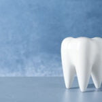 A white ceramic tooth model stands on a smooth, light blue surface with a textured blue background. The tooth model has a glossy finish and is centrally placed in the image.