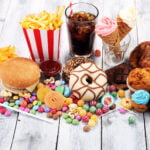 An assortment of various junk foods and sweets on a white wooden surface. The collection includes a hamburger, a container of French fries with a cup of ketchup, a glass of cola with a straw, potato chips, popcorn, colorful candies, lollipops, a chocolate bar, donuts, muffins, croissants, and ice cream cones. The arrangement is colorful and visually appealing, showcasing a variety of indulgent snacks and desserts.