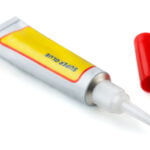 A tube of super glue with a yellow label and red text, lying on its side with the red cap removed. The nozzle is visible, and the label reads "SUPER GLUE" in blue letters. The background is a clean white surface.
