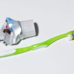 A green toothbrush with multicolored bristles lies next to a squeezed, partially used tube of toothpaste with the cap on, set against a plain white background. The toothpaste tube appears to be metallic, with visible creases from being squeezed.