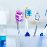 Four toothbrushes in different colors (pink, orange, blue, and grey) are placed in a frosted cup. In the background, there is a bottle of blue mouthwash with a white cap. The setting appears to be a bathroom or a dental care area, with a soft focus on the background elements.