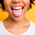Close-up of a woman with curly hair smiling and sticking out her tongue. She is wearing a white shirt and is set against a bright yellow background. The image focuses on her mouth, showcasing her healthy teeth and playful expression.