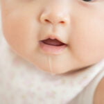 A close-up of a baby's face showing the mouth slightly open with a small line of drool coming out. The baby is wearing a light-colored bib with a floral pattern.