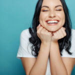 A woman with dark hair smiling brightly with her eyes closed, both hands resting under her chin. She is wearing a white t-shirt and is set against a teal background. Her expression is joyful and delighted.