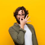 A man with curly hair and glasses, wearing an olive green shirt and white t-shirt, is pinching his nose with a disgusted expression, standing against a bright yellow background.