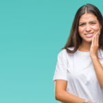 A young woman with long dark hair wearing a white t-shirt holds her hand to her cheek, wincing in pain, indicating a toothache or dental discomfort. The background is a solid teal color.
