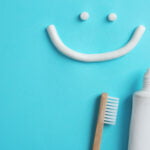 A smiley face is created using toothpaste on a blue background, with the eyes as small dots and the mouth as a curved line. Below the smiley face, there is an open tube of toothpaste with some paste coming out, and a bamboo toothbrush placed next to it.